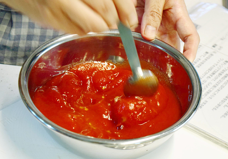 ニンニクが色づき香りが立ってきたら、缶詰のトマトをつぶしてから＝写真＝汁ごと鍋に入れ、塩ひとつまみを加えて中火で１０～１５分煮込む。最後に塩・こしょうで味を調える。